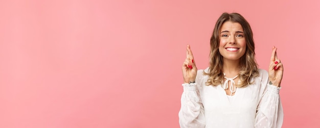 Retrato de primer plano de una hermosa chica rubia esperanzada que pide un deseo anticipando resultados importantes, cruza los dedos, buena suerte y sonríe mientras se siente un poco nervioso, usa un vestido blanco de fondo rosa