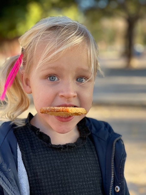 Retrato de primer plano de una hermosa chica rubia que parece divertida con una galleta en los dientes.