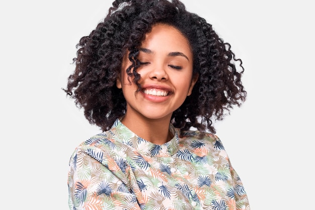 Retrato de primer plano de una hermosa joven de piel oscura vestida con una camisa con motivos florales que se siente feliz durante una conversación con una amiga afroamericana sonriendo ampliamente posando sobre una pared blanca
