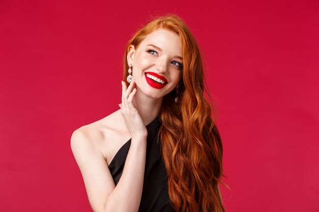 Retrato de primer plano de la hermosa y lujosa y sofisticada modelo femenina joven pelirroja rica asistir a la semana de la moda o fiesta en vestido de noche negro, aretes y lápiz labial rojo, sonriendo mirar lejos soñador