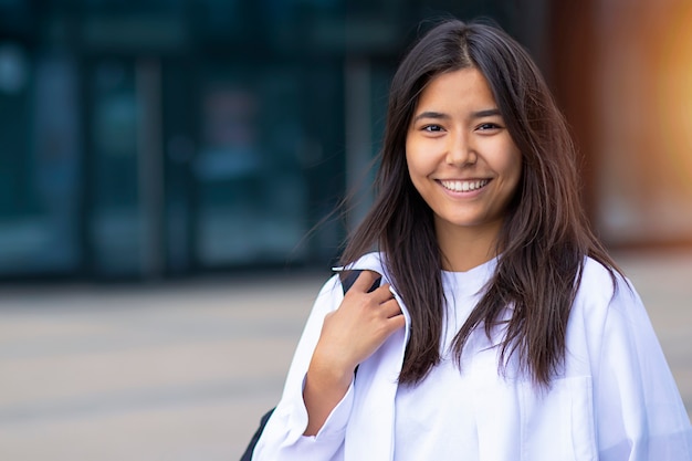 Retrato, primer plano, hermosa mujer asiática agradable o niña sonriente con mochila. Señora alegre al aire libre. Estudiante afuera con camisa blanca. Copie el espacio.