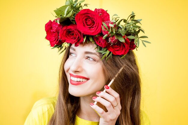 Retrato de primer plano de una hermosa mujer con corona de rosas rojas hablando con el teléfono sobre el fondo amarillo