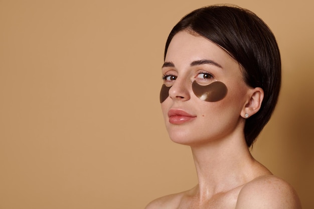 Retrato de primer plano de una hermosa mujer joven de cabello oscuro con piel fresca y saludable que se aplica parches para los ojos alisantes de colágeno médico debajo de los ojos contra el fondo de color beige con espacio publicitario de copia