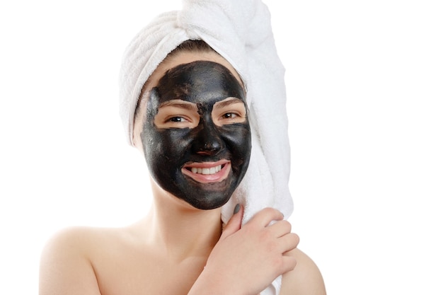 Retrato de primer plano hermosa mujer con máscara facial negra sobre fondo blanco, niña con una toalla blanca en la cabeza, sonrisa satisfecha y feliz