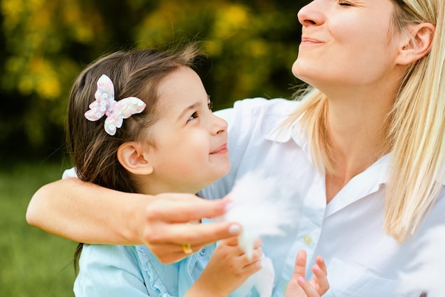 Retrato de primer plano de una hija feliz comiendo algodón de azúcar con su madre sentada en el césped en el parque Mujer joven disfrutando el tiempo junto con su lindo hijo al aire libre Día de la madre