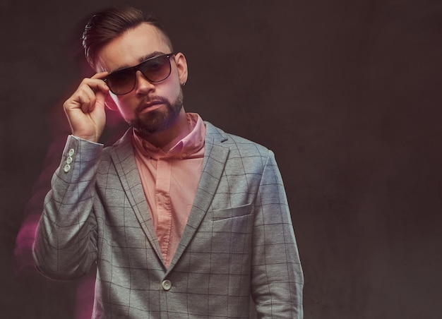 Retrato de primer plano de un hombre barbudo elegante y confiado con peinado y gafas de sol en un traje gris y camisa rosa, posando en un estudio. Aislado en un fondo gris.