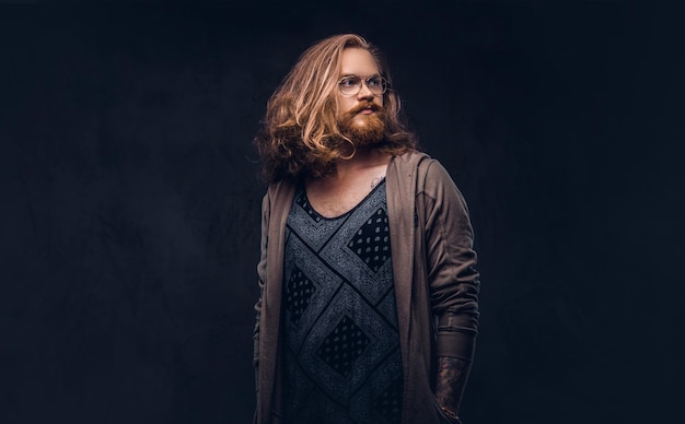 Retrato de primer plano de un hombre hipster pelirrojo con cabello largo y exuberante y barba completa vestido con ropa informal de pie en un estudio, mirando hacia otro lado. Aislado en un fondo oscuro.