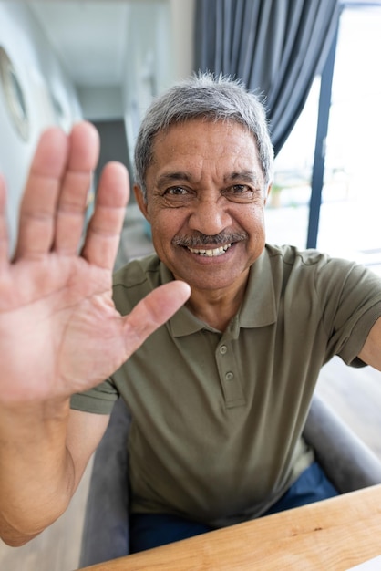 Retrato en primer plano de un hombre mayor biracial alegre agitando la mano mientras habla por video llamada. Sin alteración, estilo de vida, feliz, gesto, comunicación, concepto de hogar y jubilación.