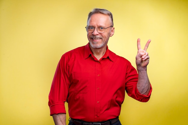 Retrato de primer plano de un hombre de negocios sonriente mayor sosteniendo un signo de paz victoria dos aislado sobre fondo amarillo Emoción positiva expresiones faciales símbolos actitud comunicación Éxito en la vida