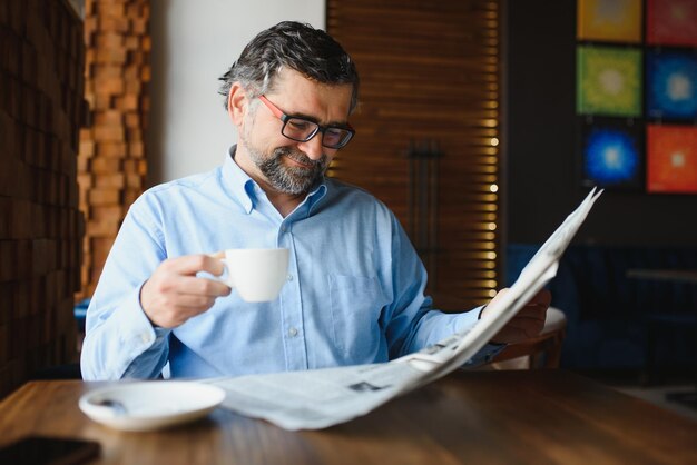 Retrato de primer plano de un hombre serio y guapo leyendo el periódico tomando un descanso para tomar café y sentado en la mesa