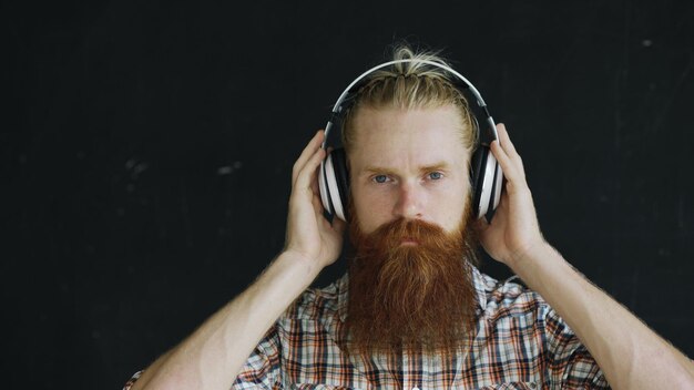 Retrato de primer plano de un joven barbudo con auriculares escuchando música y mirando a la cámara sonriendo