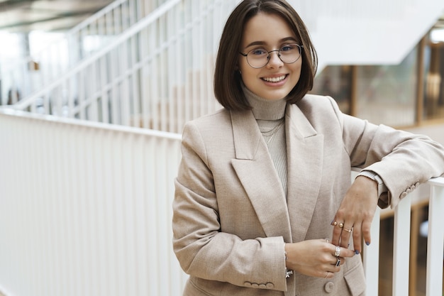 Retrato de primer plano de una joven empresaria inteligente y profesional, una chica que comienza su carrera en la empresa, se apoya en la escalera y sonríe a la cámara, usa gafas de chaqueta beige, habla con compañeros de trabajo