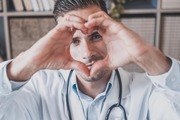 Foto retrato en primer plano de un joven enfermero caucásico o médico de cabecera sonriente en uniforme médico blanco muestra corazón amor gesto de la mano joven médico feliz muestra apoyo y cuidado a los pacientes o clientes en el hospitalxa