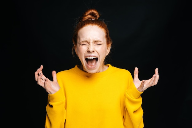 Retrato de primer plano de una joven enojada emocionada con suéter amarillo gritando con los ojos cerrados sobre fondo negro aislado Modelo de dama pelirroja bonita que muestra emocionalmente el espacio de copia de expresiones faciales