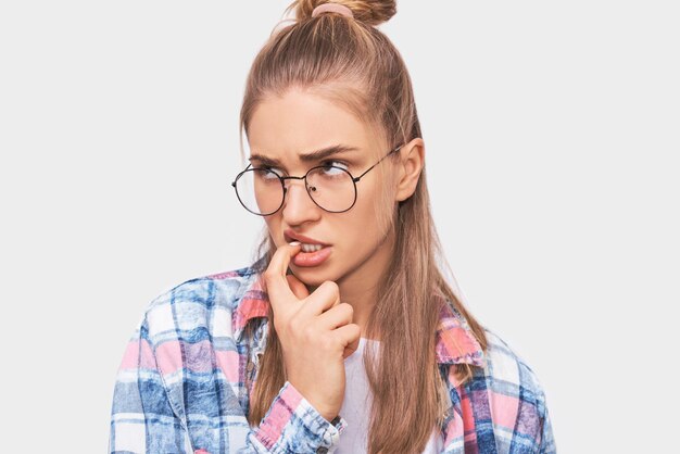 Retrato de primer plano de una joven estudiante seria mirando a un lado con el dedo índice en los labios usando ropa informal y anteojos redondos transparentes Mujer bonita rubia posando en el estudio