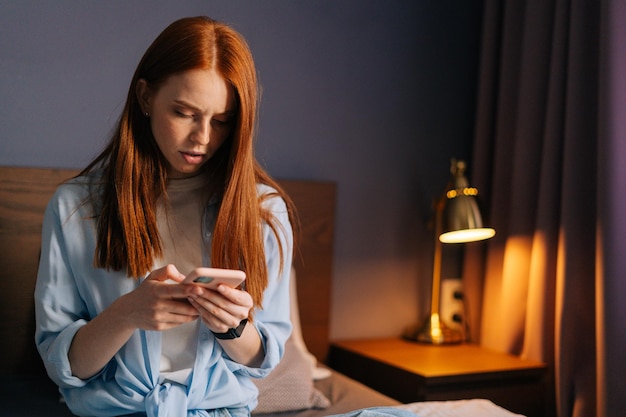 Retrato de primer plano de una joven seria escribiendo un mensaje en el teléfono móvil en el dormitorio. Mujer sonriente con ropa de casa chateando en línea por teléfono celular. Linda dama tocando el celular en casa.