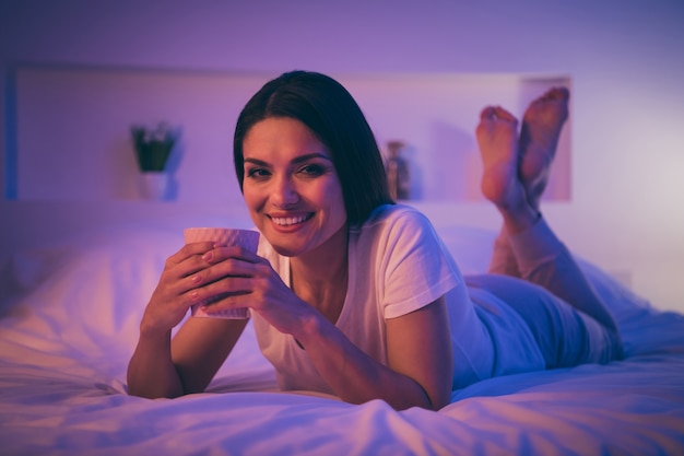 Retrato de primer plano de linda mujer alegre acostada en la cama disfrutando de la bebida de descanso