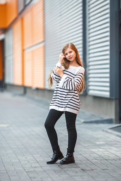 Retrato de primer plano de una linda niña modelo sonriente con chaqueta a rayas posando cerca de una pared de rayas corrugadas de color naranja gris con una mochila roja