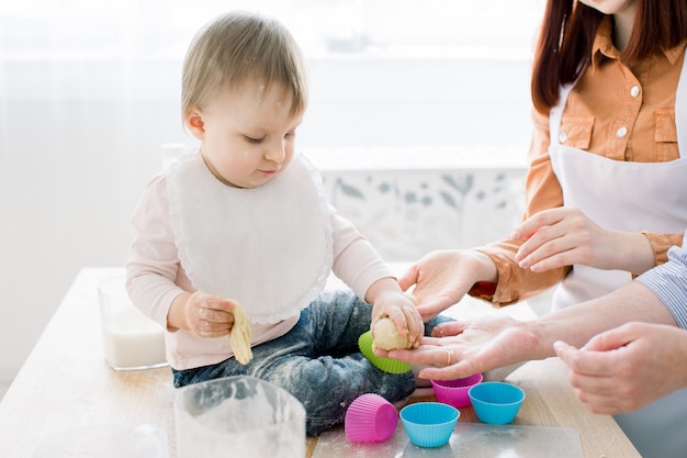 Retrato de primer plano de linda niña sentada en la mesa y jugando con la masa para hornear muffins en formas de colores. Madre y abuela trabajando con masa. Concepto del día de la madre