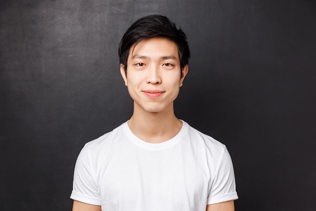 Retrato de primer plano de un modelo masculino asiático joven sonriente de aspecto amable en camiseta blanca, mirando a cámara y sonriendo, de pie, concepto de emociones y estilo de vida de las personas