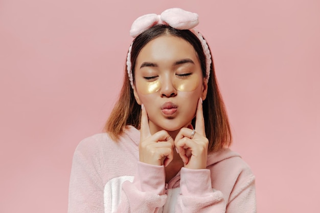 Retrato de primer plano de mujer asiática de pelo corto en kigurumi y diadema sopla beso y posa con los ojos cerrados sobre fondo rosa