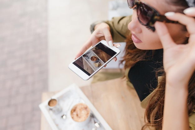 Retrato de primer plano de mujer concentrada con manicura blanca haciendo imagen de su almuerzo. Fotografía aérea de una niña con gafas de sol sosteniendo un teléfono inteligente sobre un café y un croissant.