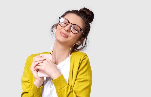 Retrato de primer plano de una mujer joven y bonita con anteojos que disfruta de la música favorita de los auriculares usando un teléfono inteligente Mujer joven caucásica con piel brillante viste blusa amarilla y camiseta blanca Gente