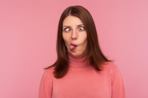 Retrato de primer plano de una mujer loca divertida que cruza los ojos y muestra la lengua, divirtiéndose haciendo una expresión de cara tonta, engañando a la cara. Disparo de estudio interior aislado sobre fondo rosa