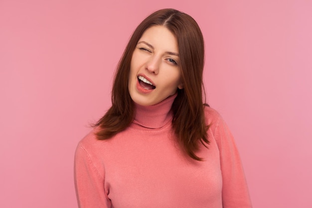 Foto retrato de primer plano de mujer morena divertida coqueta en suéter rosa guiñando un ojo mirando a la cámara animando con guiño disparo de estudio interior aislado sobre fondo rosa