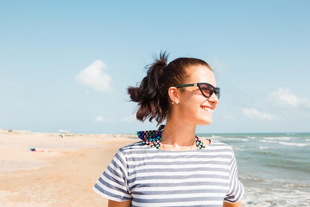 Retrato primer plano mujer morena feliz en un vestido a rayas con gafas de sol junto al mar mira las olas risas y sonrisas Turismo playa vacaciones brisa viajes viajes