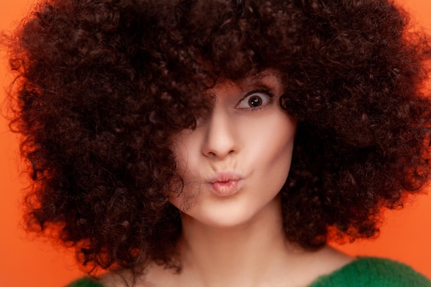 Retrato de primer plano de mujer con peinado afro mirando a la cámara con labios de puchero con piel perfecta y cabello esponjoso anunciando salón de belleza Estudio interior aislado sobre fondo naranja