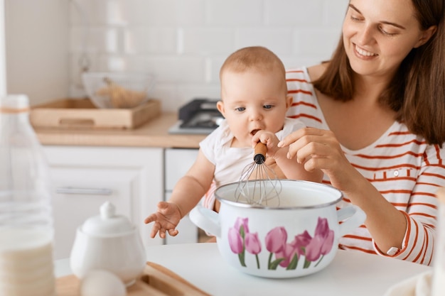 Foto retrato de primer plano de una mujer positiva sonriente con camisa a rayas horneando junto con su hija pequeña sentada en la mesa con olla y whist y mezclando ingredientes para la masa