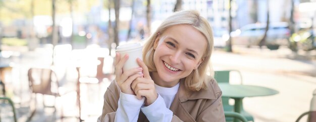 Retrato en primer plano de una mujer rubia sonriente estudiante universitaria sosteniendo una taza de café calentándola