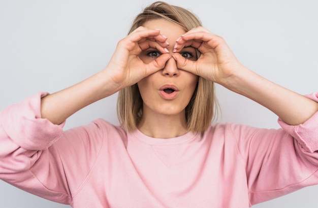 Retrato de primer plano de una mujer rubia sorprendida con un suéter casual rosa sosteniendo sus manos en sus ojos como si mirara a través de binoculares o anteojos Mujer sorprendida contra el fondo de la pared blanca del estudio