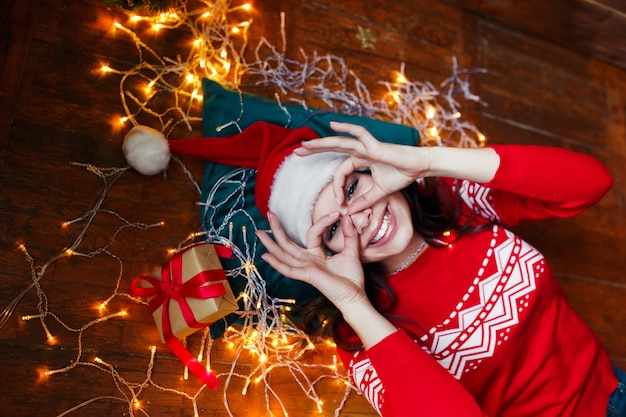 Retrato de primer plano de mujer con sombrero de Santa envuelto en luces de Navidad divirtiéndose y acostado en el piso