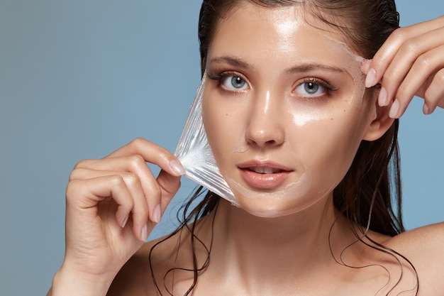 Retrato de primer plano de niña bonita quitándose la máscara facial transparente aislada en azul