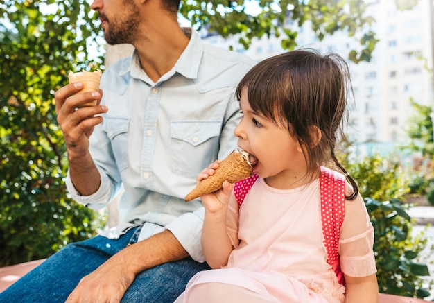 Retrato de primer plano de una niña feliz y linda sentada con papá en la calle de la ciudad y comiendo helado al aire libre Niña divertida y padre se divierten afuera Buena relación entre papá e hija