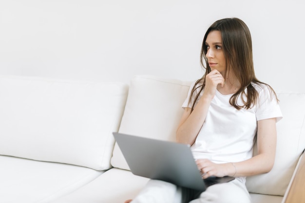 Retrato de primer plano de niña preguntando mirando a otro lado sentado en un sofá suave con un portátil blanco en la sala de estar luminosa. Mujer pensativa está escribiendo texto en el cuaderno