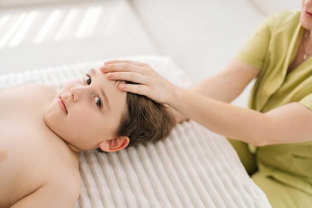 Retrato en primer plano de un niño de cinco años que recibe tratamiento osteopático o quiropráctico en una clínica pediátrica mirando a la cámara En primer plano de una osteópata haciendo terapia fisiológica del cráneo