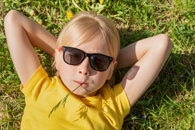 Retrato en primer plano de un niño rubio con una camiseta amarilla y gafas de sol acostado en la hierba Un niño descansa sobre diente de león archivado con las manos detrás de la cabeza