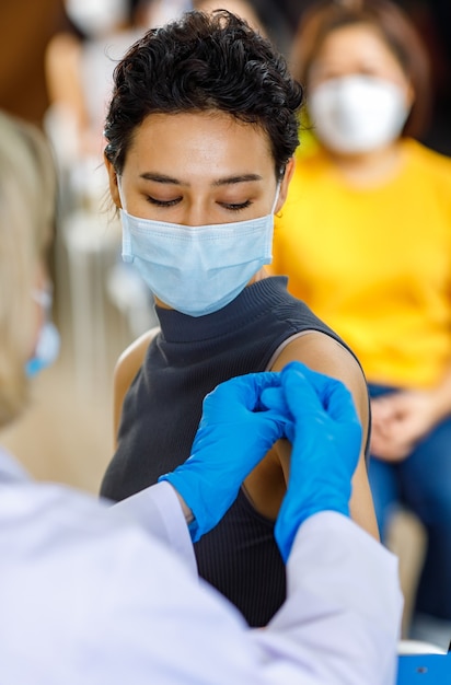 Retrato de primer plano de una paciente usa mascarilla, sentarse, mirar a la cámara mientras está en la cola de vacunación, recibir la inyección de la vacuna disparada por el médico en bata de laboratorio blanca y guantes de goma azules.