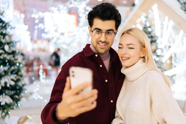 Retrato de primer plano de una pareja joven feliz tomando una foto selfie en un teléfono móvil de pie en el salón del centro comercial Celebra en Nochebuena