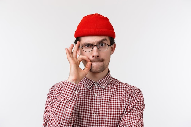 Retrato de primer plano de un serio joven caucásico con bigote, usar anteojos y gorro, sellar los labios con el gesto de los dedos como si estuviera ocultando el secreto, promete no hablar de eso, pararse