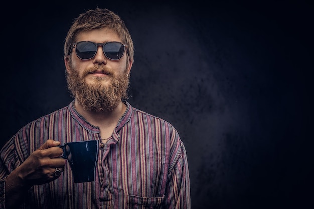 Retrato de primer plano de un tipo hipster con gafas de sol vestido con una camisa pasada de moda sostiene una taza de café. Aislado en un fondo oscuro.