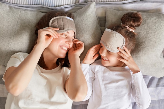 Retrato de primera vista de una mujer de cabello castaño con su hija con máscaras para dormir acostada en la cama mirándose con una sonrisa y amor despertándose en la mañana del fin de semana