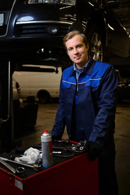 Retrato profesional de un apuesto hombre caucásico, mecánico de automóviles, técnico, ingeniero de automóviles parado cerca de una caja con un conjunto de herramientas para reparar un automóvil levantado en un polipasto en el taller de reparación