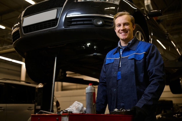 Retrato profesional de un apuesto hombre caucásico, mecánico de automóviles, técnico, ingeniero de automóviles, sonriendo a la cámara de pie cerca de una caja con herramientas para reparar automóviles en el taller de reparación