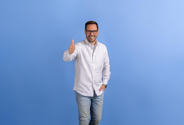 Retrato de un profesional masculino positivo con la mano en el bolsillo mostrando el pulgar hacia arriba en un fondo azul