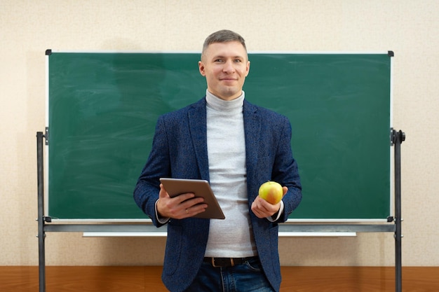 Retrato de un profesor sonriente con una tableta y una manzana verde en el aula