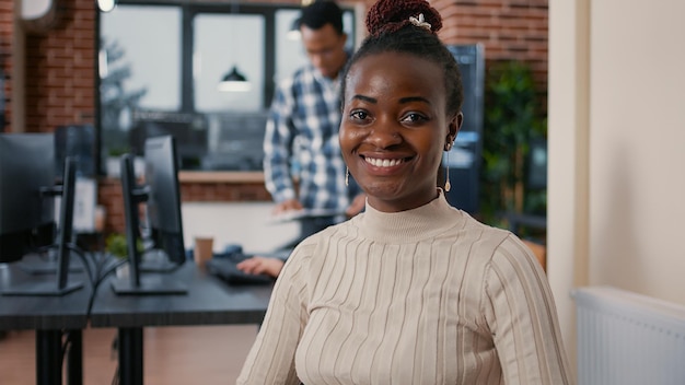 Retrato de un programador afroamericano sentado codificando en una laptop mirando hacia arriba y sonriendo a la cámara. Codificador que usa una computadora portátil junto a un equipo mixto en una agencia de desarrollo de software.